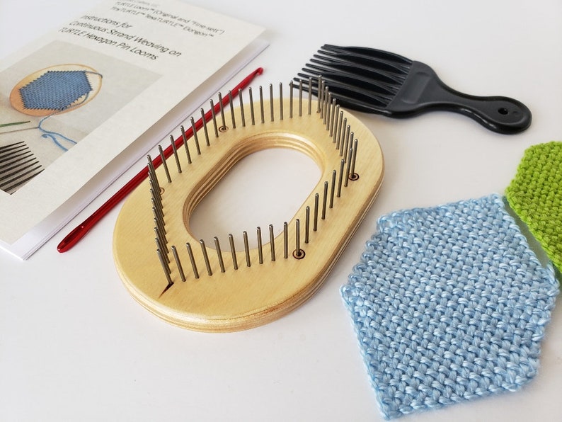 May include: A wooden knitting loom with a rounded shape and a central opening. The loom has rows of metal pegs and is accompanied by a red crochet hook, a black comb, and two knitted squares in blue and green.