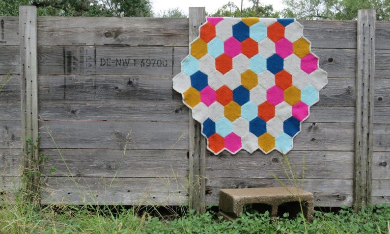 May include: A colorful crocheted blanket with a geometric pattern of hexagons in shades of pink, blue, orange, yellow, and white. The blanket is hanging on a wooden fence.