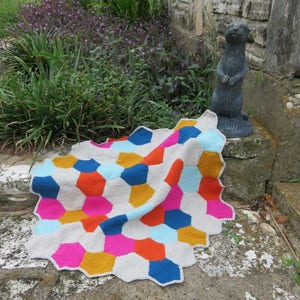 May include: A colorful crocheted blanket with a geometric pattern of hexagons in shades of blue, pink, orange, and yellow. The blanket is folded and draped over a stone step.