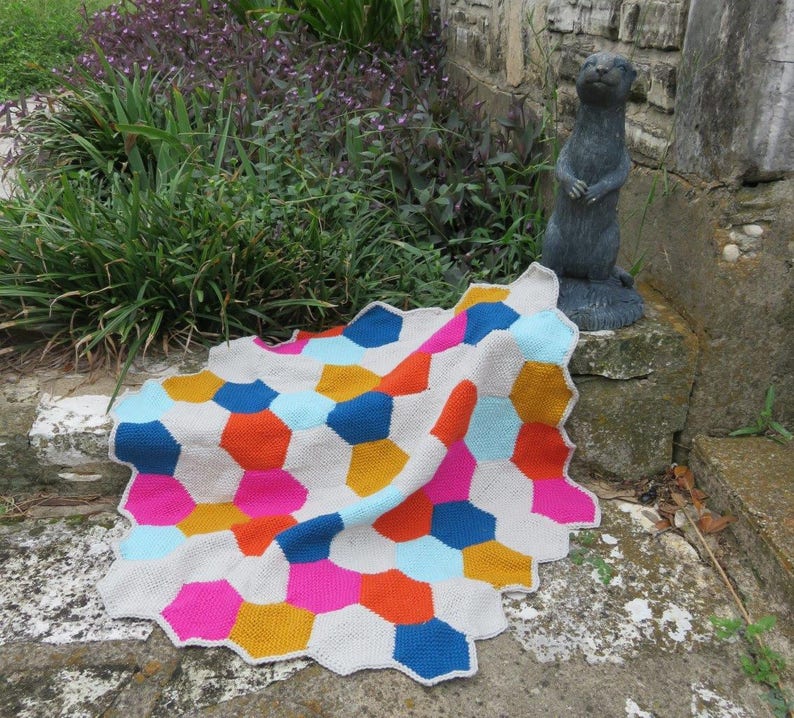 May include: A colorful crocheted blanket with a geometric pattern of hexagons in shades of blue, pink, orange, and yellow. The blanket is folded and draped over a stone step.