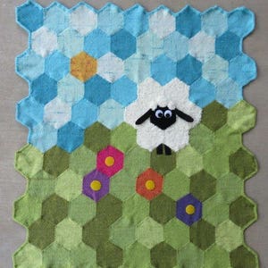 May include: A knitted baby blanket with a hexagonal pattern in shades of green, blue, and white. A white sheep with black eyes and a black nose is in the center of the blanket. There are colorful flowers around the sheep.