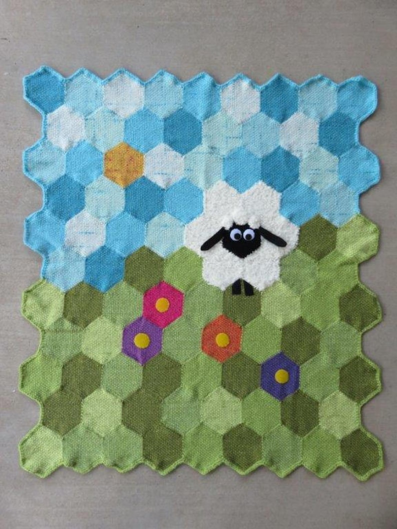 May include: A knitted baby blanket with a hexagonal pattern in shades of green, blue, and white. A white sheep with black eyes and a black nose is in the center of the blanket. There are colorful flowers around the sheep.