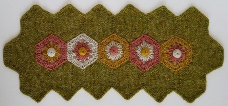May include: A green crocheted table runner with a scalloped edge and five crocheted flower designs in shades of pink, white, and yellow.
