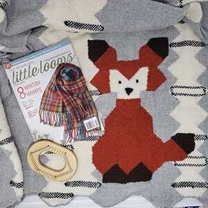 May include: A close-up of a grey and white woven blanket with a red fox design. The blanket is on a table with a magazine titled "little looms" and a wooden loom tool.