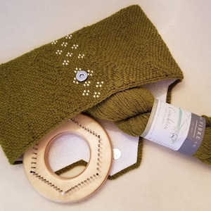 May include: A green knitted cowl with a white stitch pattern, a ball of green yarn, a label that reads "Fibre Co.  Cotswold" and a wooden knitting loom.