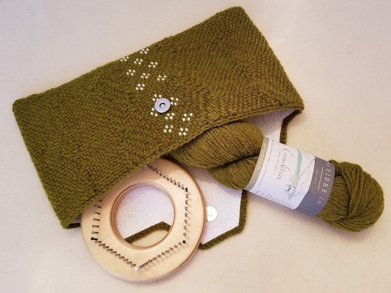 May include: A green knitted cowl with a white stitch pattern, a ball of green yarn, a label that reads "Fibre Co.  Cotswold" and a wooden knitting loom.