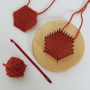 May include: A close-up of a red yarn weaving project on a wooden loom. The yarn is woven into a heart shape and is being worked on with a red crochet hook. There is also a ball of red yarn and a finished woven heart shape.