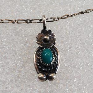May include: Silver-toned pendant necklace featuring a turquoise-colored stone. The pendant has a floral design at the top and a decorative border around the stone. The necklace chain is delicate and has a lobster clasp.