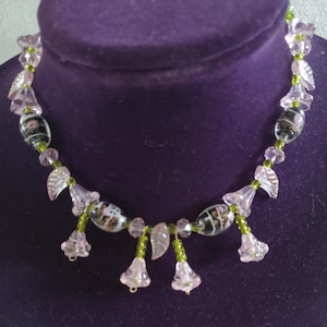 May include: A delicate necklace featuring clear, pink, and black glass beads. The necklace has a floral design with pink flower pendants and green accents. The beads are strung on a green cord, creating a unique and elegant piece of jewelry.