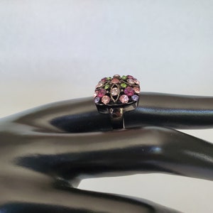 May include: A black ring with a square-shaped setting featuring a colorful design of pink, purple, green, and clear rhinestones.