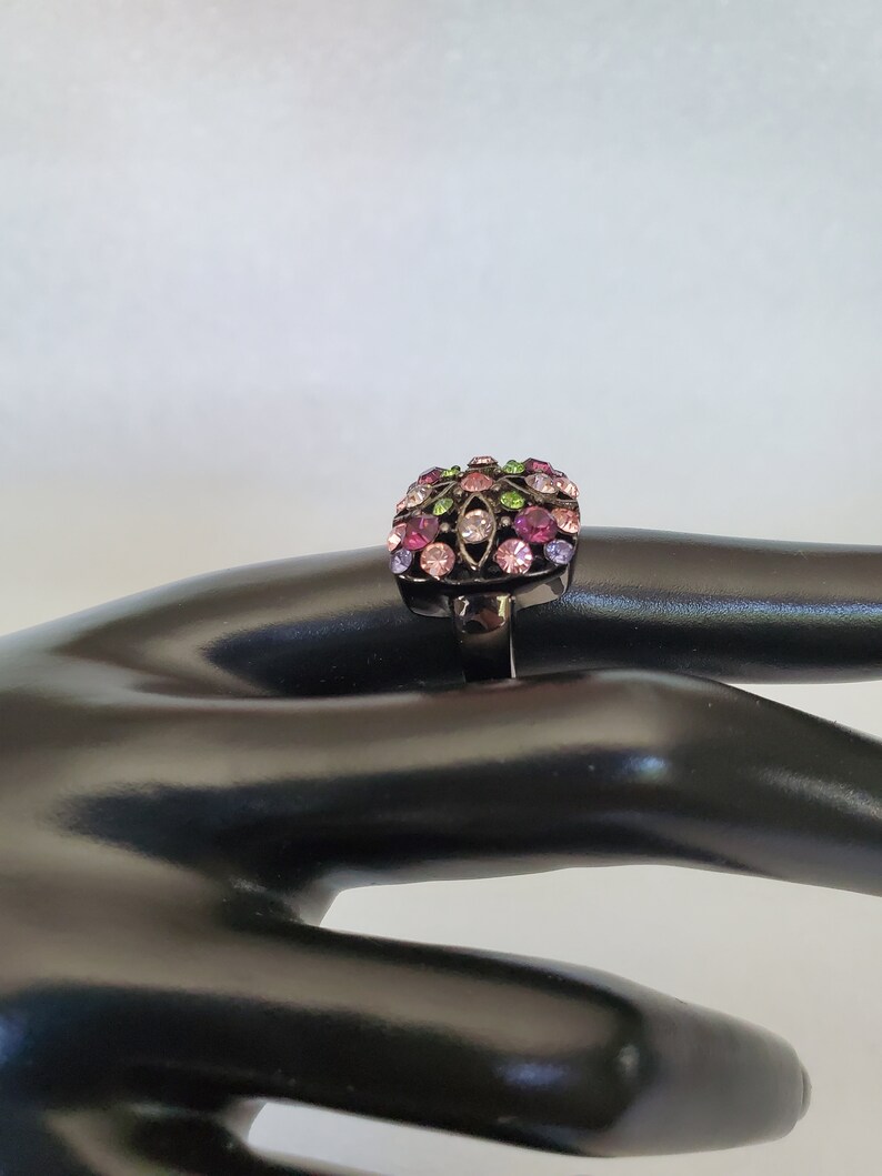 May include: A black ring with a square-shaped setting featuring a colorful design of pink, purple, green, and clear rhinestones.