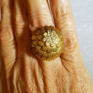 May include: A gold ring with a large, round, flower-shaped setting. The setting is adorned with clear, round rhinestones.