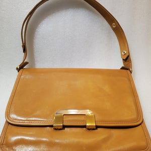 May include: A tan leather handbag with a rectangular shape and a flap closure. The bag features a gold-colored metal clasp and a matching shoulder strap. The bag is approximately 12 inches wide and 8 inches tall.