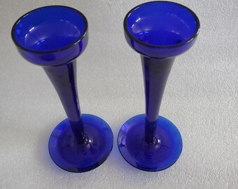 May include: Two cobalt blue glass vases with a flared top and a slender stem. The vases have a round base and a dark rim around the top. The vases are likely for displaying flowers or other decorative items.