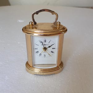 May include: A gold-toned Howard Miller quartz carriage clock with a white face, black Roman numerals, and black hands. The clock features a polished metal frame with vertical fluted side columns and a decorative arched handle on the top.