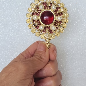 May include: A gold-tone compact mirror with a red jewel in the center surrounded by faux pearls and rhinestones. The mirror has a decorative floral design.