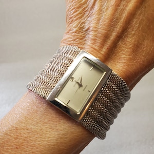 May include: A silver watch with a rectangular face and a mesh band. The watch face has a white background with black hour and minute hands and black Roman numeral hour markers. The watch face also has the words "Anne Klein" and "Diamond" printed on it.
