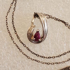 May include: Silver pendant necklace featuring a teardrop-shaped setting with a pear-cut ruby gemstone. The pendant has a diamond-studded accent and hangs from a delicate chain. The clasp is marked with the text "TREBOR PARIS".