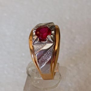 May include: A two-tone ring featuring a central round-cut red gemstone set in a silver-colored mounting. The band is crafted from polished yellow gold, while the center section displays a textured, silver-colored finish with geometric detailing.
