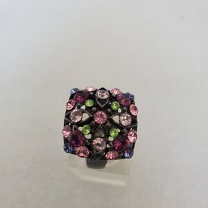 May include: A square-shaped ring with a black metal setting and a floral design. The ring is adorned with pink, purple, and green rhinestones.