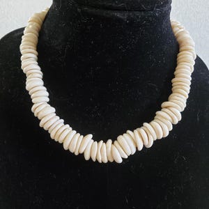 May include: A white shell necklace made of small, oval-shaped shells. The necklace is on a black velvet display stand.