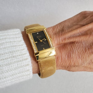 May include: Gold-toned rectangular watch with a black face and a mesh band. The watch face has the text "KJL" and "QUARTZ". The watch is adorned with small, clear crystals along the edges of the face.
