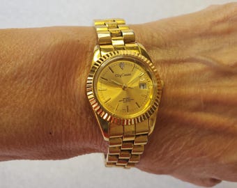 May include: A gold-tone wristwatch with a gold-tone metal band. The watch face is round and has a gold-tone bezel with a small diamond at the top. The watch face is gold-tone and has black markings. The watch face also has the text "Olga Cassini" and "Swiss Made Quartz" printed on it.