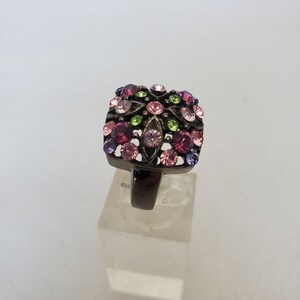 May include: A square-shaped ring with a black metal band and a cluster of colorful rhinestones in shades of pink, purple, green, and clear.