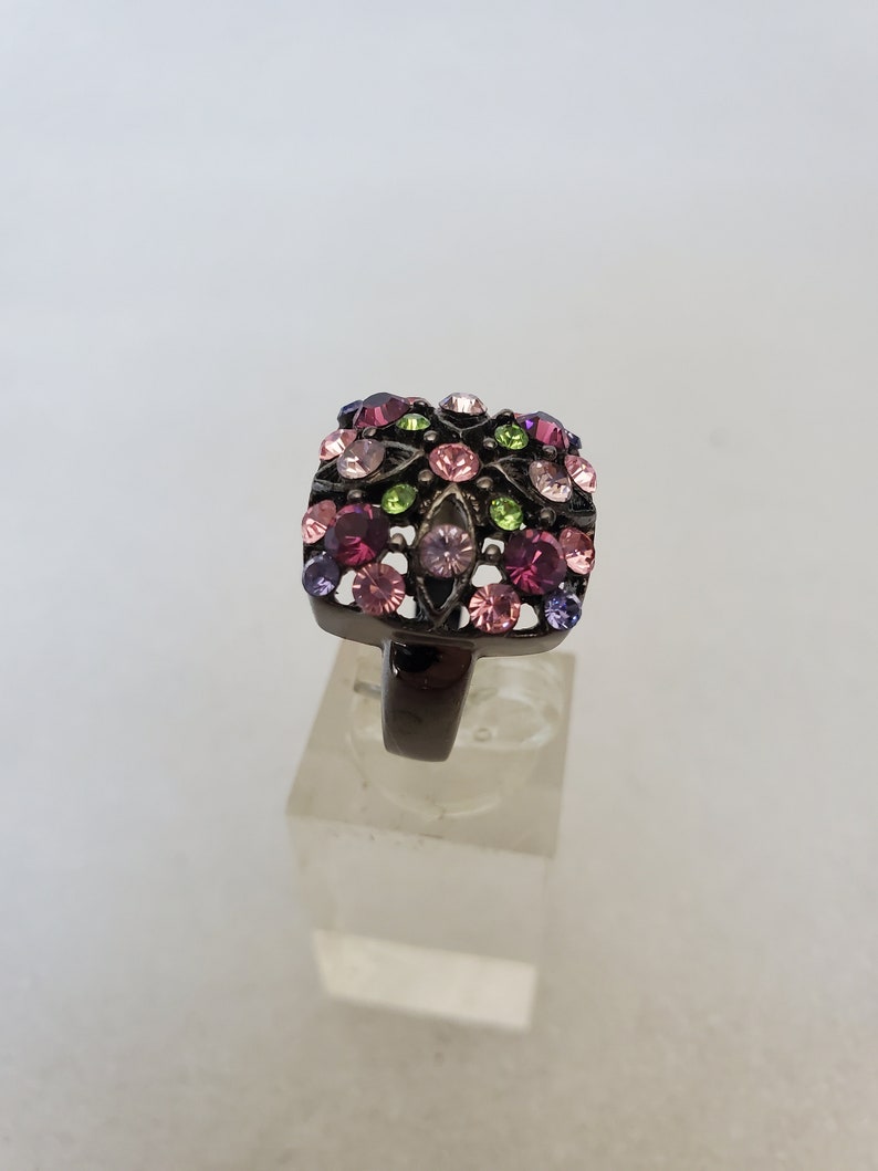 May include: A square-shaped ring with a black metal band and a cluster of colorful rhinestones in shades of pink, purple, green, and clear.