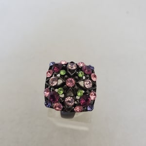 May include: A square-shaped ring with a black metal setting and a floral design. The ring is adorned with pink, purple, and green rhinestones.