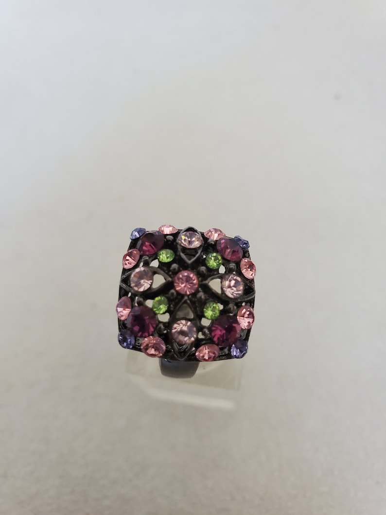 May include: A square-shaped ring with a black metal setting and a floral design. The ring is adorned with pink, purple, and green rhinestones.