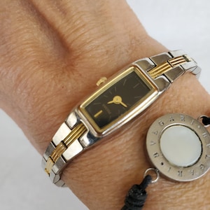 May include: A rectangular Pulsar wristwatch with a black face, gold-tone bezel, and a two-tone silver and gold metal link band. Beside it is a circular Bvlgari charm featuring a white center inlay on a thin black cord with a knotted detail.