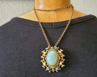 May include: A vintage gold-tone necklace featuring a large oval pendant. The pendant has a speckled turquoise center stone, surrounded by small turquoise and white accents. The necklace is displayed on a black top.