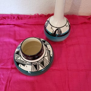May include: Two ceramic vases with a Southwestern design. The larger vase is white with a blue and black geometric pattern. The smaller vase is white with a green and black geometric pattern.
