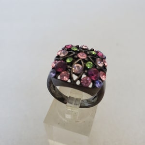 May include: A black ring with a square-shaped setting adorned with colorful rhinestones in shades of pink, purple, green, and clear.