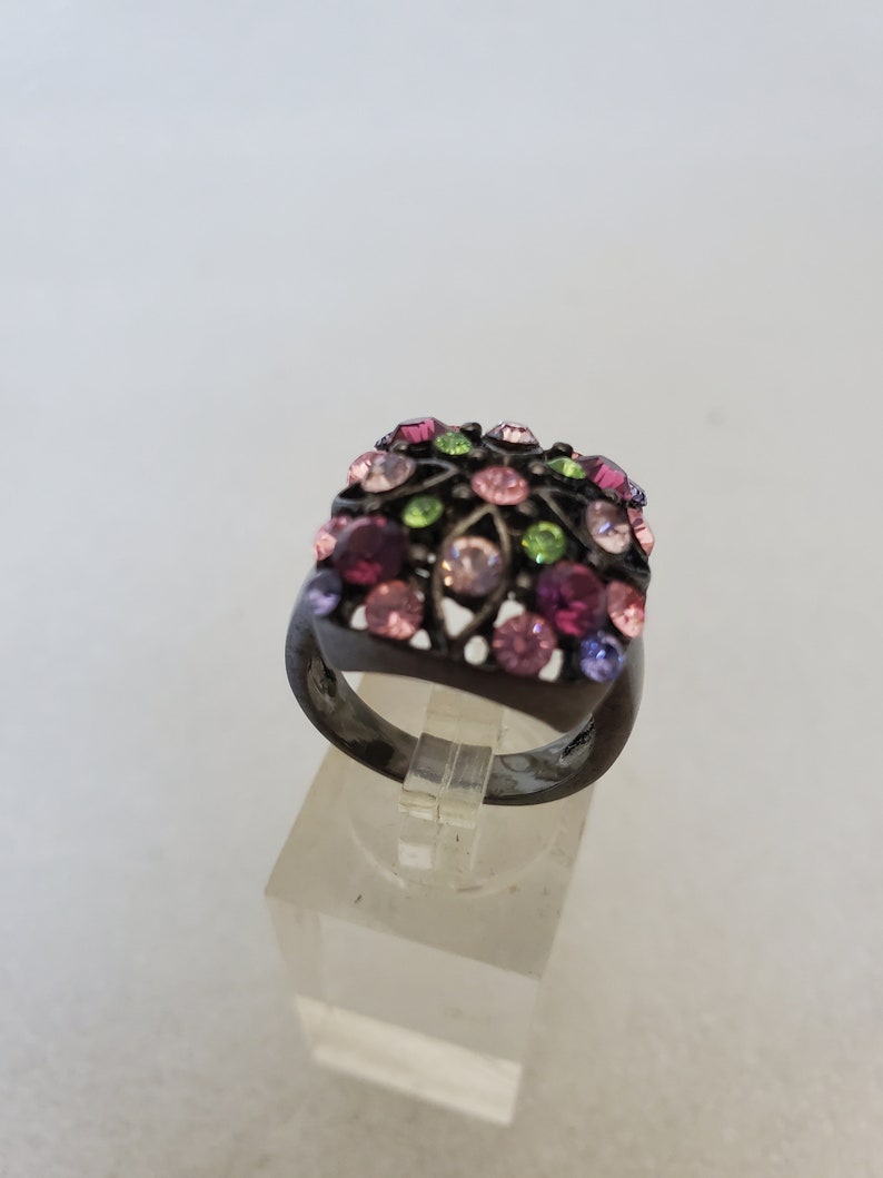 May include: A black ring with a square-shaped setting adorned with colorful rhinestones in shades of pink, purple, green, and clear.
