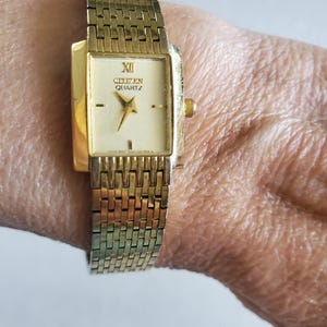 May include: A gold-toned Citizen Quartz watch with a rectangular face and a matching linked band. The watch face is cream-colored with black Roman numerals and hands. The watch is fastened on a wrist.