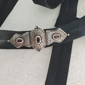 May include: A black leather choker necklace with a decorative silver-toned centerpiece. The centerpiece features three sections with floral designs and three oval-shaped red gemstones. The choker has a black ribbon.