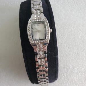May include: Silver-toned rectangular wristwatch with a silver dial and crystal accents. The watch has a matching silver band with crystal embellishments. The watch is displayed on a black surface.