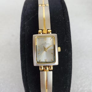 May include: Anne Klein watch with a rectangular silver-tone face and a silver and gold-tone band. The watch face has gold hands and Roman numeral hour markers. The band is composed of rectangular silver links with gold accents.
