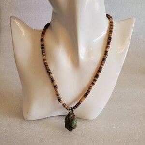 May include: A necklace with a pendant and a beaded chain. The pendant is a rough, green stone. The chain is made of small, flat, brown and tan beads. The necklace is displayed on a white mannequin.