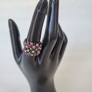 May include: A black ring with a square-shaped setting filled with colorful rhinestones. The rhinestones are pink, purple, green, and clear.