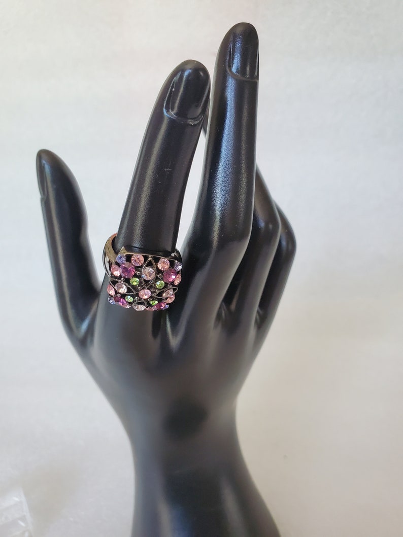 May include: A black ring with a square-shaped setting filled with colorful rhinestones. The rhinestones are pink, purple, green, and clear.