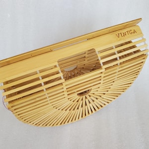 May include: A semi-circular bamboo handbag featuring a slatted construction held together by thin white connectors. The bag has a flat top edge with the word VINTGA engraved on the right side and a central cutout handle integrated into the design.