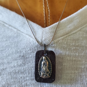 May include: A silver chain necklace with a rectangular pendant. The pendant is dark brown with a gold-colored religious figure in an oval frame. The necklace is worn over a white t-shirt.