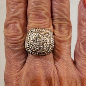 May include: A silver-toned ring with a square face, encrusted with numerous small, clear stones. The ring has a rope-like border around the square face. The ring is on a hand with visible wrinkles.