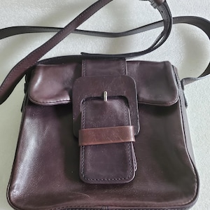 May include: A dark brown leather crossbody bag with a large rectangular buckle closure. The bag has a flap and a long, adjustable strap. The buckle is a lighter shade of brown than the bag itself.
