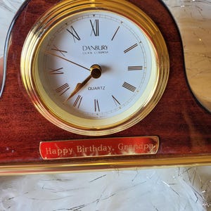 May include: A wooden clock with a gold trim and a white face. The clock face has black Roman numeral hour markers and black hands. The clock has a gold plaque with the inscription "Happy Birthday, Grandpa".