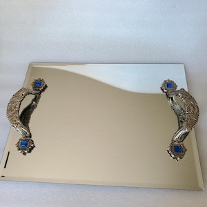 May include: A silver mirrored serving tray with ornate handles and blue gemstone accents. The tray is rectangular and has a reflective surface.