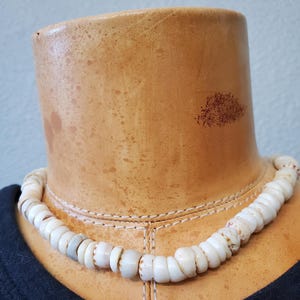 May include: A white shell necklace with a natural, organic design. The necklace is composed of rounded, off-white shells, strung together to form a choker. The shells have subtle variations in color and texture, creating a unique, earthy aesthetic.